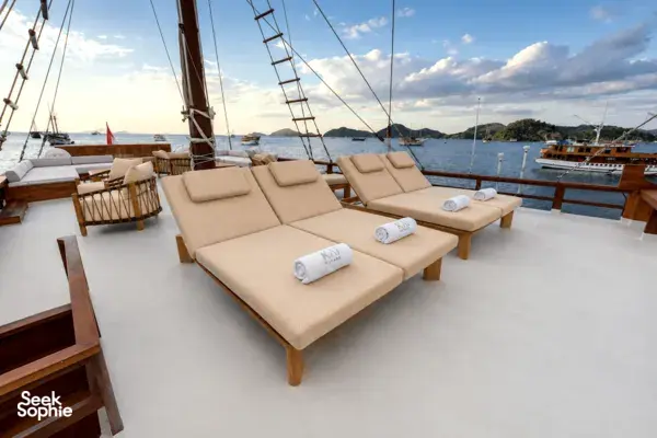 Komodo Island phinisi upper deck laid out with cushioned sun loungers and folded towels, framed by sail spars and sea views