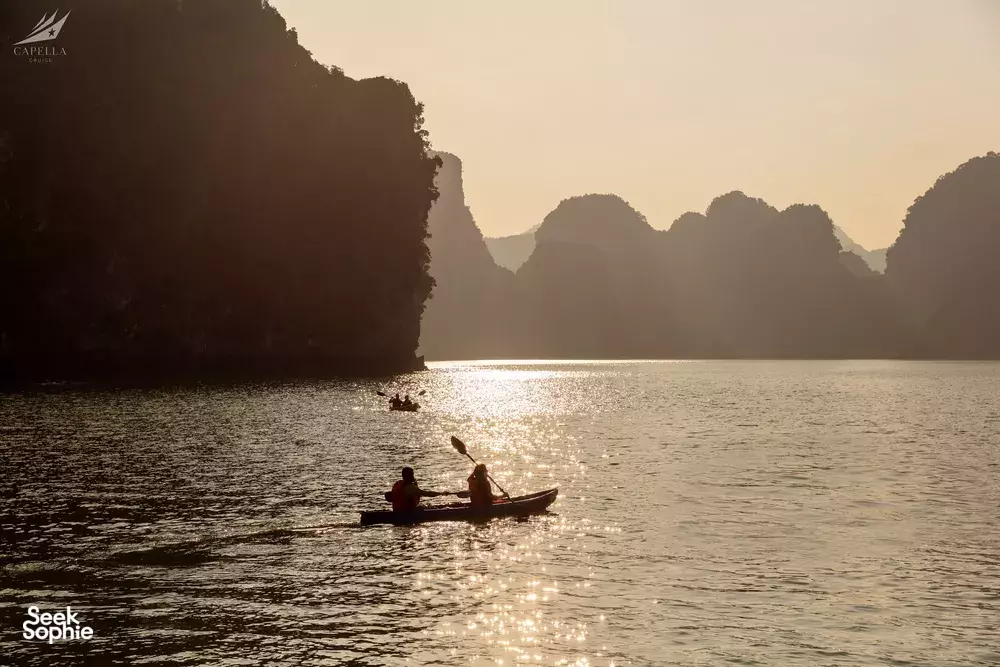 Silhouette of a kayaker paddling across Halong Bay’s calm waters at golden hour between limestone cliffs—Lyra Cruise Collection