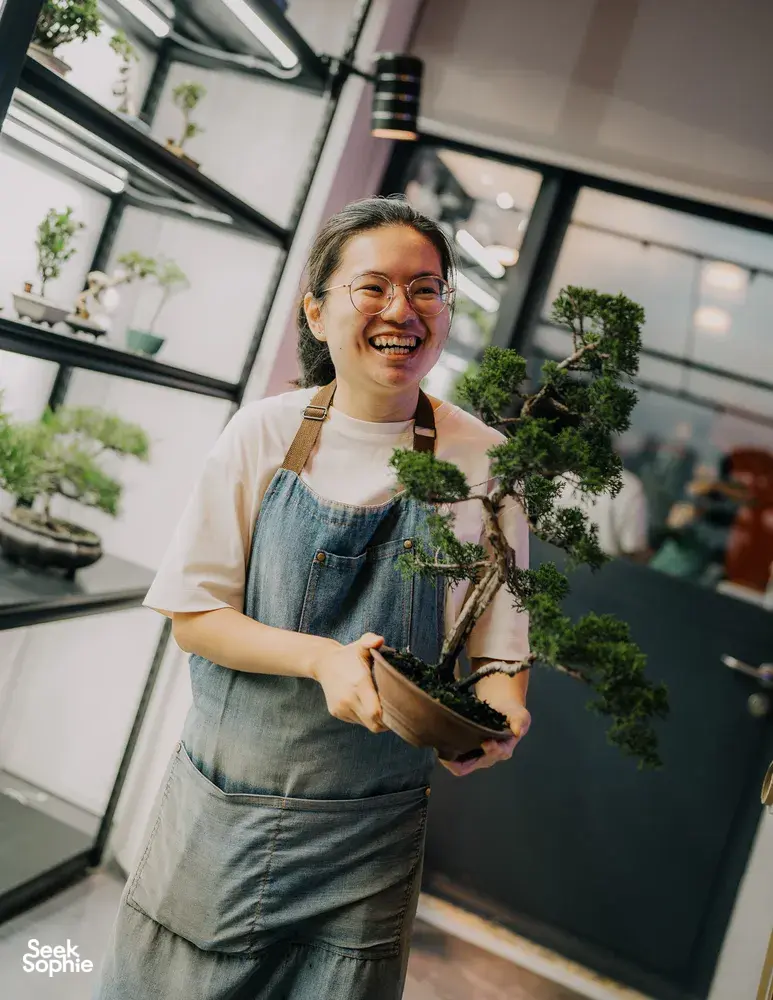 Smiling woman lifting a small tropical bonsai in a dark pot inside Bonsai Inc in Singapore, with shelves and trees behind