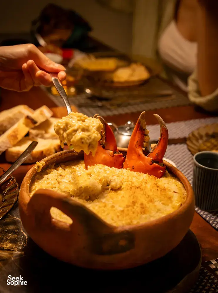 Hand lifting orange prawns from a clay bowl of creamy stew, with bread on the side during a 5-course Patagonian-inspired dinner in Singapore