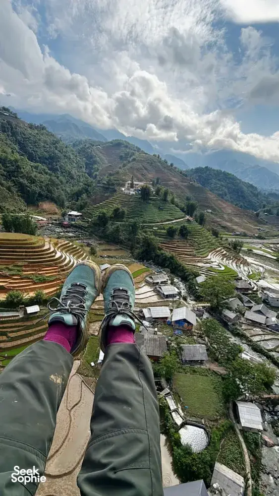 Paragliding passenger view over terraced rice valleys and mountain village paths far below, with clouds overhead, above Vietnam's Sapa