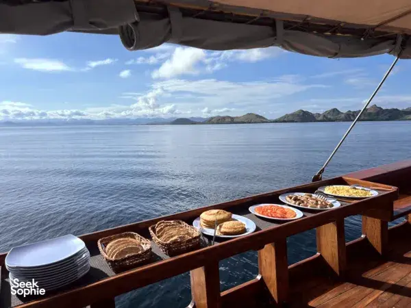 Breakfast on the open boat deck with plates of bread, fruit and drinks as the voyage heads through calm water near Labuan Bajo