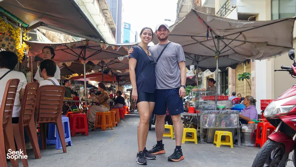 在胡志明市热闹的街市小巷里,两位旅行者撑着伞站着,参加 Jackfruit Adventure 的单车一日行程