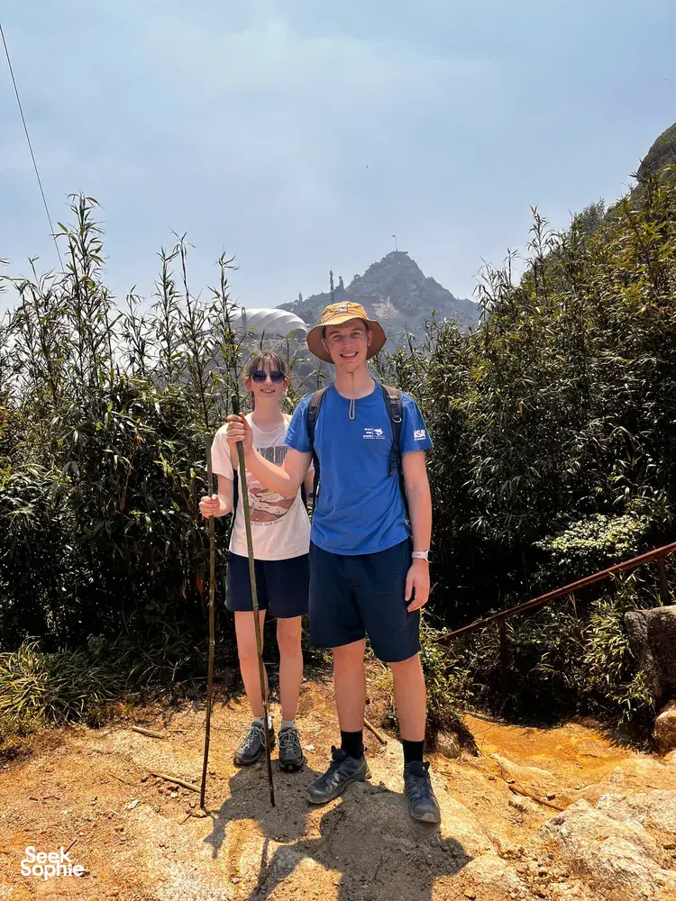 Two hikers pause on a narrow trail through tall grass near Mt Fansipan in Sapa, trekking with poles on a 1-day eco trek