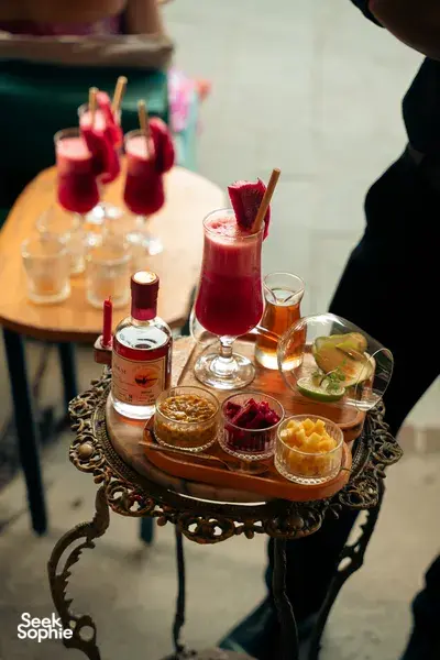 Red cocktail in a stem glass with watermelon garnish on a decorative serving tray with spirit bottle and appetisers, Hoi An