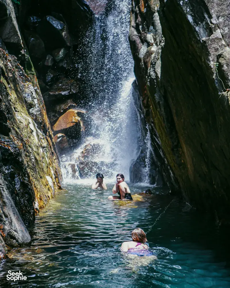 People wading in a narrow gorge pool as water cascades down the rocks on a Hoi An secret waterfall stop