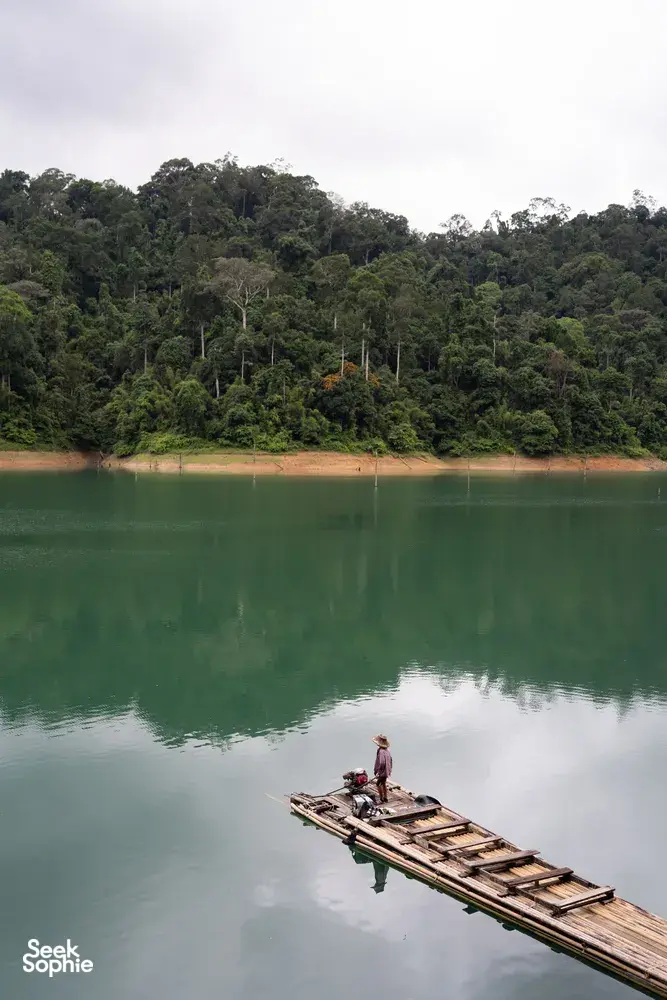 Travellers ride a bamboo raft on glassy green water with forested karst hills in the distance at Cheow Lan Lake