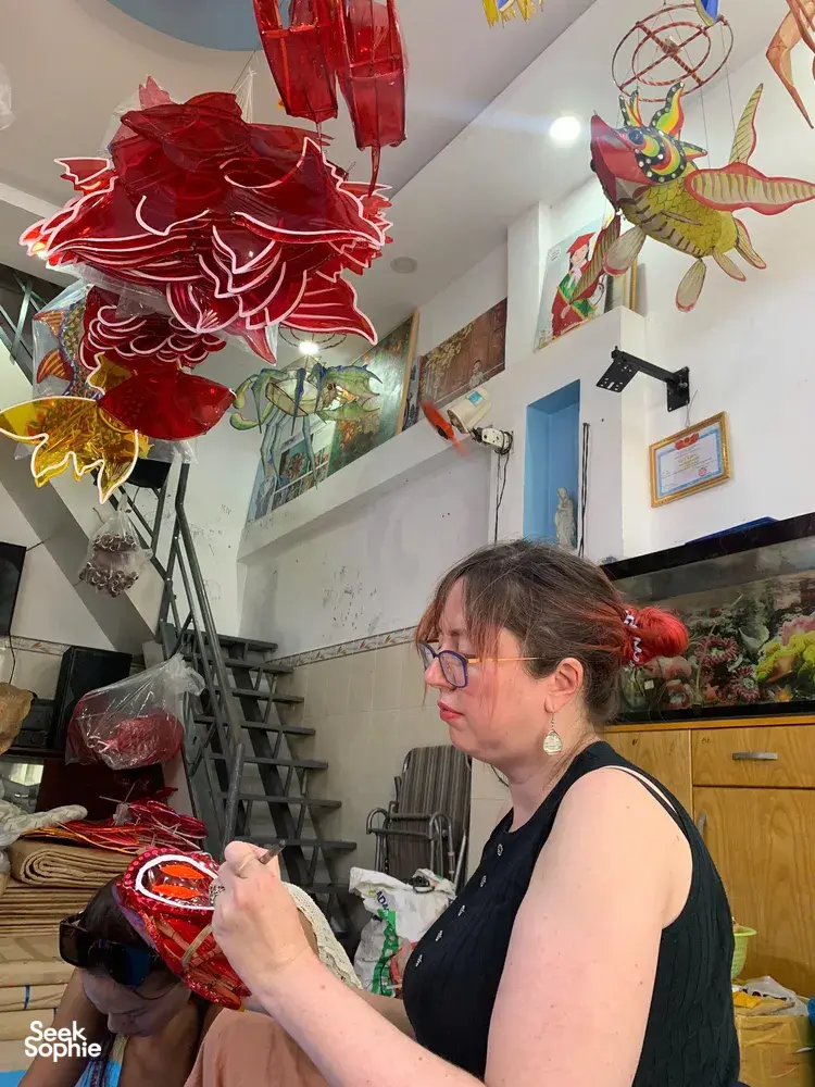 Woman assembling and painting red paper lanterns while other colourful hanging lanterns fill a Ho Chi Minh City craft studio