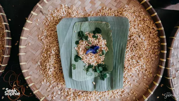 Grains and sprigs of herbs scattered on a square green leaf plate set in a woven tray, presented as Bali foodie meal