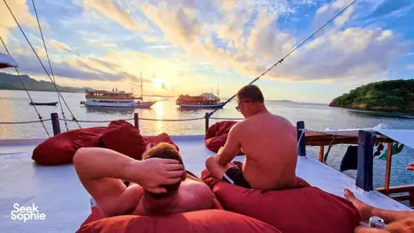Two travellers lounging on the deck at golden hour with the boat and glassy sea behind them on a Komodo liveaboard Labuan Bajo