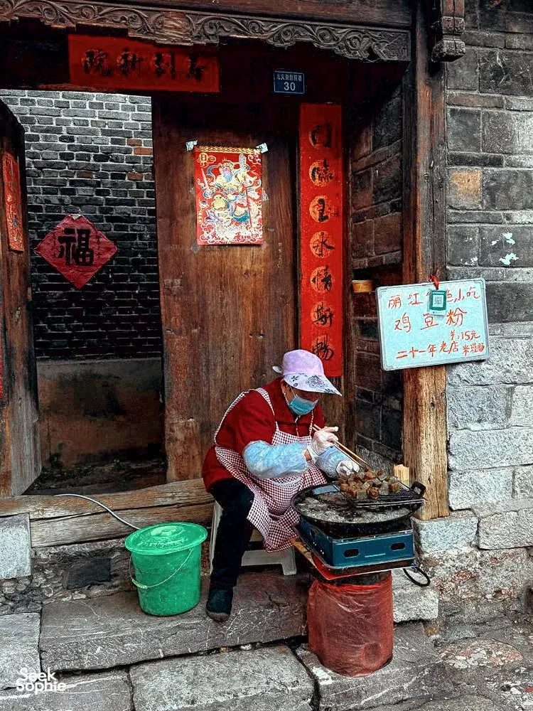 Street food cook tending a small stove on a stone step in Lijiang Old Town, beside hanging Chinese signs and a wooden door
