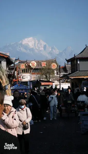 Crowded narrow market street in Lijiang old town with traditional shops, Chinese signboards, and snow-dusted mountains in view