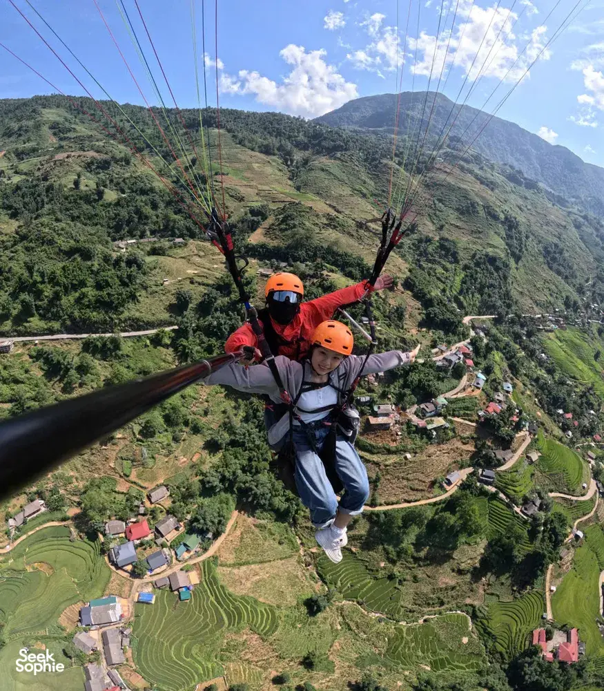 Tandem paragliding with an orange-helmeted pilot and passenger soaring above green mountain slopes and village farmland in Sapa