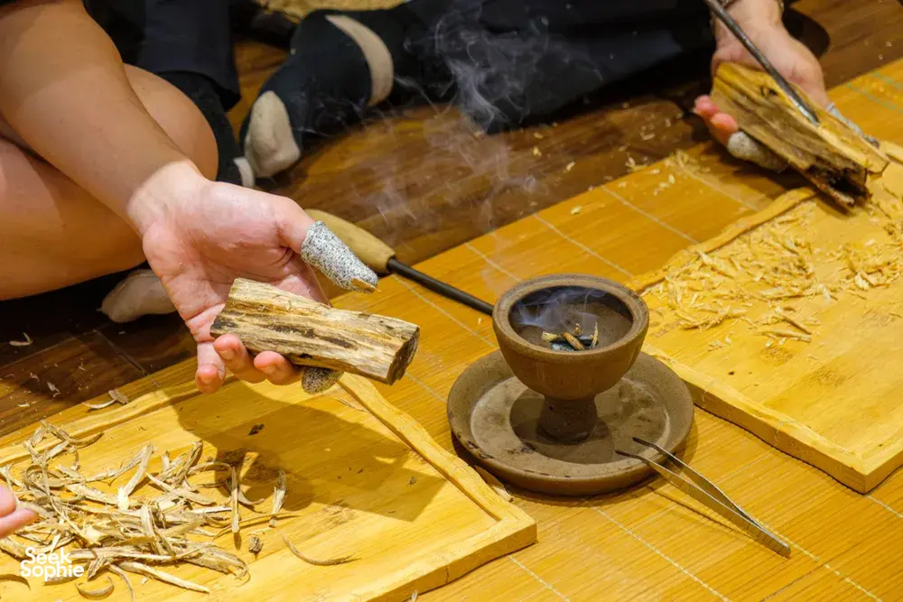Hand holds a carved agarwood stick as smoke rises from a small burner on a bamboo mat during the Hanoi tour