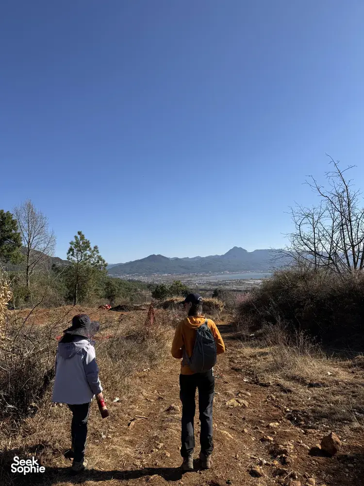 Backpackers hike a country trail in Lijiang toward a wide mountain valley, with Xintuo Ecotourism on the Yi & Naxi trek