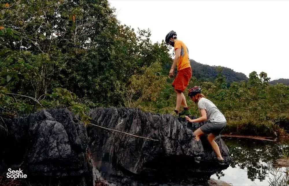 Cyclists climb a dark rock ledge by the river while a fellow rider stands nearby, surrounded by dense jungle near Kuching