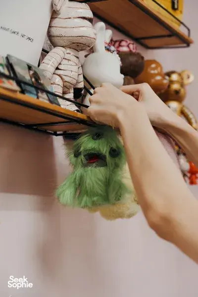 Hand placing a green ugly-cute plushie keychain on a display shelf of stuffed characters at Avant-Garde Art Space workshops in Singapore