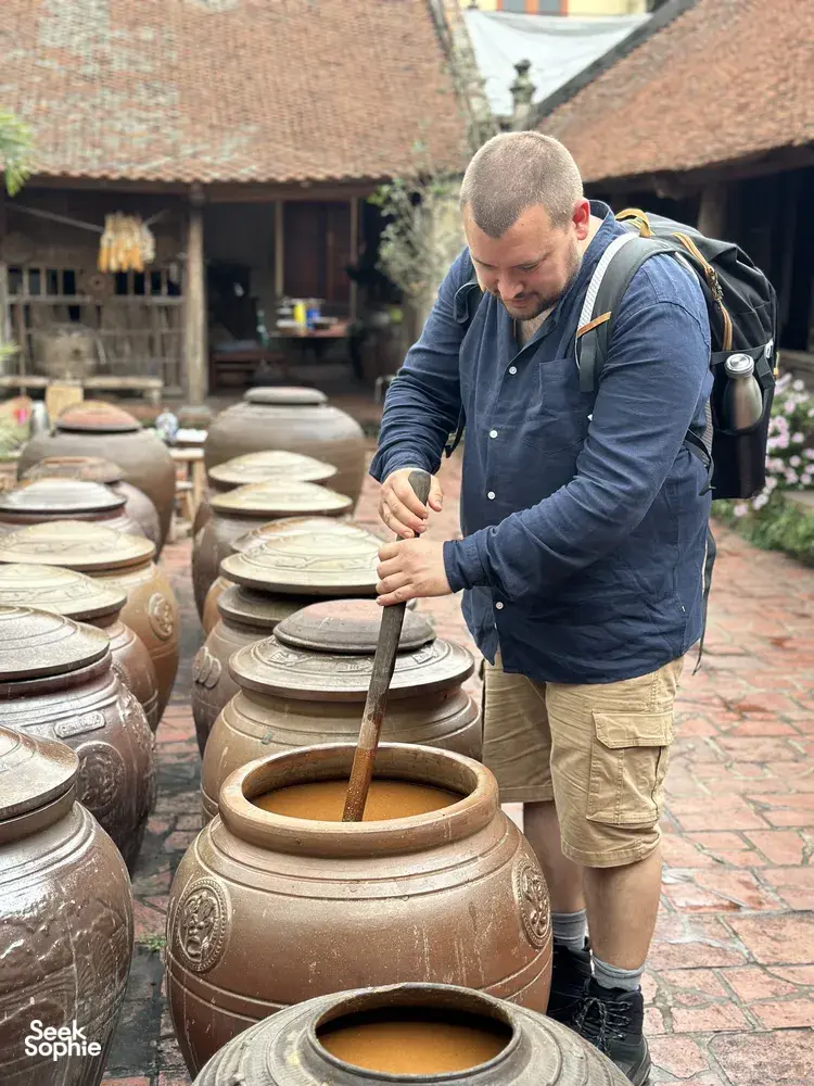 Visitor stirring liquid in a large clay jar among stacked pottery in a courtyard at Duong Lam ancient village near Hanoi during rice farming