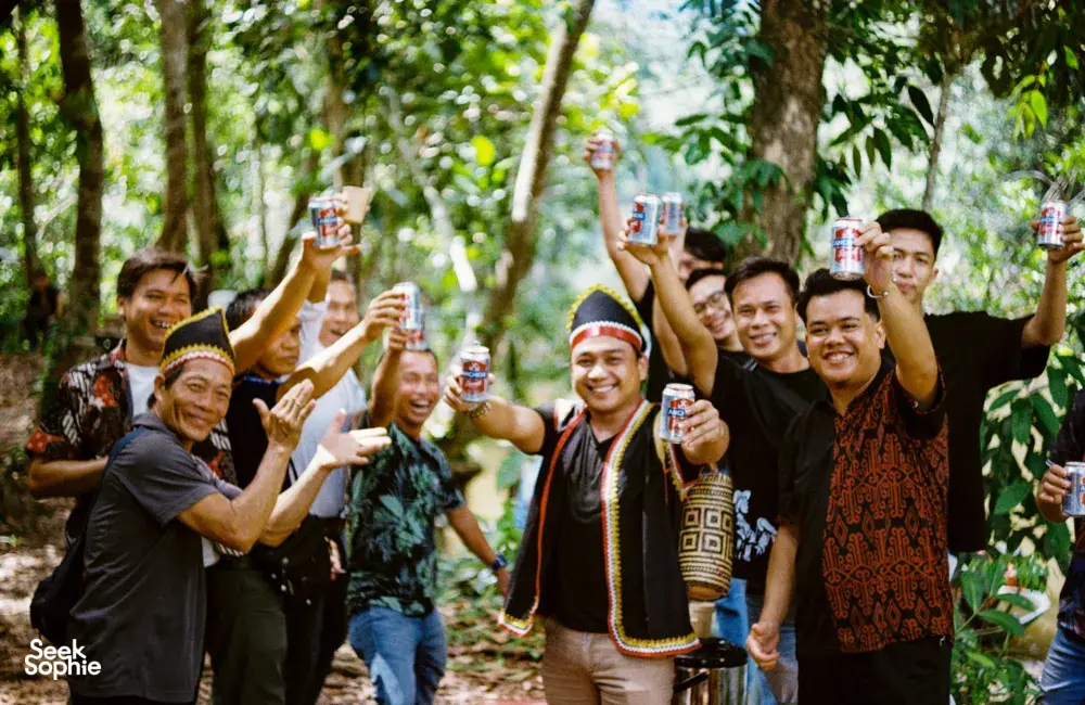 Sekumpulan jiran dan pengembara tersenyum sambil mengangkat minuman di ruang luar Ngapuh Homestead di hutan hujan Kuching