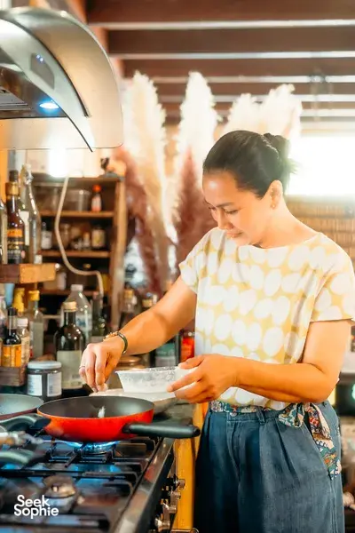 Chiang Mai home cooking class: a chef stirs food in a red pan on the gas stove, reaching for ingredients beside a spice bar.