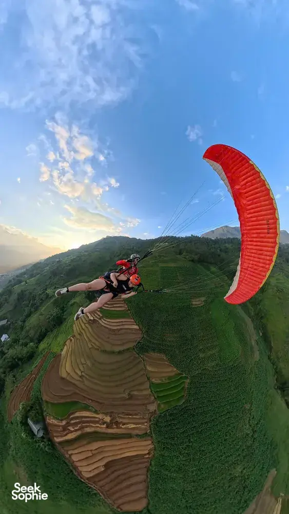 Paragliding over Sapa terraced valleys at sunset, red canopy overhead as a tandem rider floats above farmland
