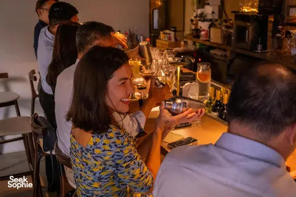 Small group gathered at the bar counter in Singapore, watching a mixologist prepare cocktails amid bottles and glassware.