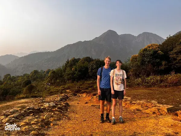 Two travellers walk along a sunlit track through woodland in Sapa, with rugged peaks towering in the distance