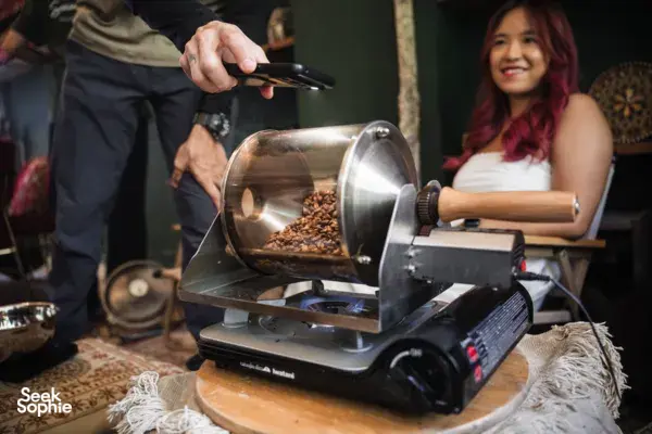 Beans being tipped into an electric coffee roaster as a woman smiles in the background during hands-on coffee roasting workshops in Singapore