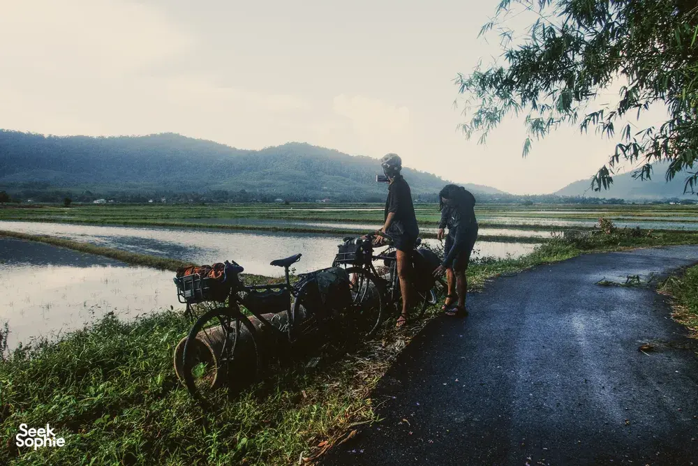 Cyclists pause beside rural fields in Langkawi on Bicycle Day Langkawi, with bikes by the water and distant hills
