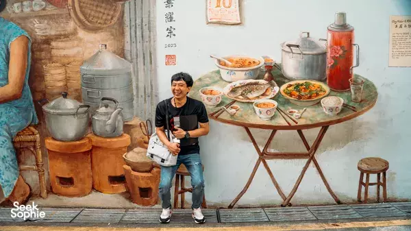 Triad Trails ex-gangster guide stands in front of a recreated hawker kitchen mural with cooking pots and a street-food table in Singapore