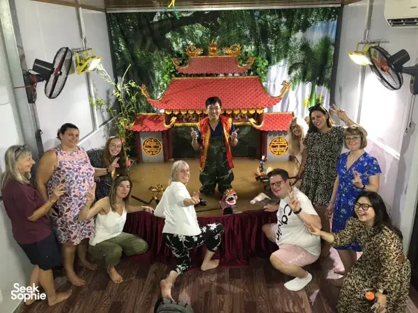 Para tamu berkumpul di sekitar panggung wayang air di Hanoi saat dalang tampil, dengan latar set beratap merah yang bersinar di belakang