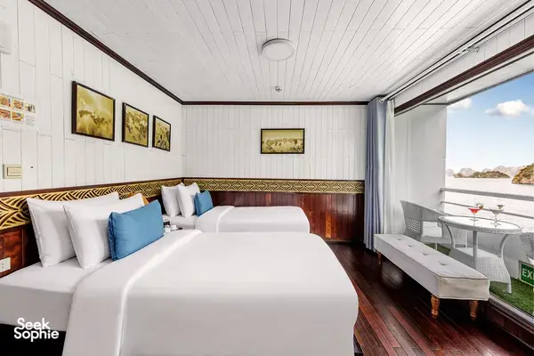 Halong Bay party cruise cabin with neatly made beds, blue pillows and dark wood floors, plus an open door to the deck