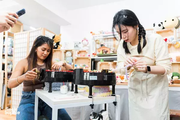 Two women at Tiny Rabbit Hole Singapore work at a pom-pom plush-making machine among colourful craft shelves — workshops in Singapore