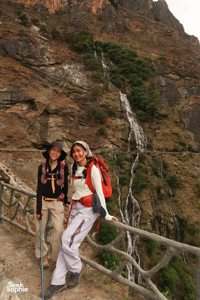 Two hikers with backpacks pause by a barrier overlooking a waterfall in Tiger Leaping Gorge near Lijiang on the 2D1N high trail