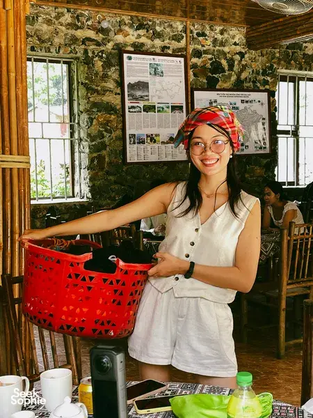 Smiling Ha Giang Expeditions host in a rustic stone room, standing by a table with travel materials and a red basket