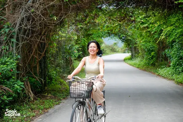 Cyclist riding along a quiet road enveloped by tropical greenery in Cat Ba, with a front basket and relaxed posture