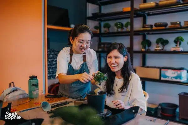 Two participants in aprons working on a tropical bonsai together at a Japanese-style workshop in Singapore, surrounded by tools