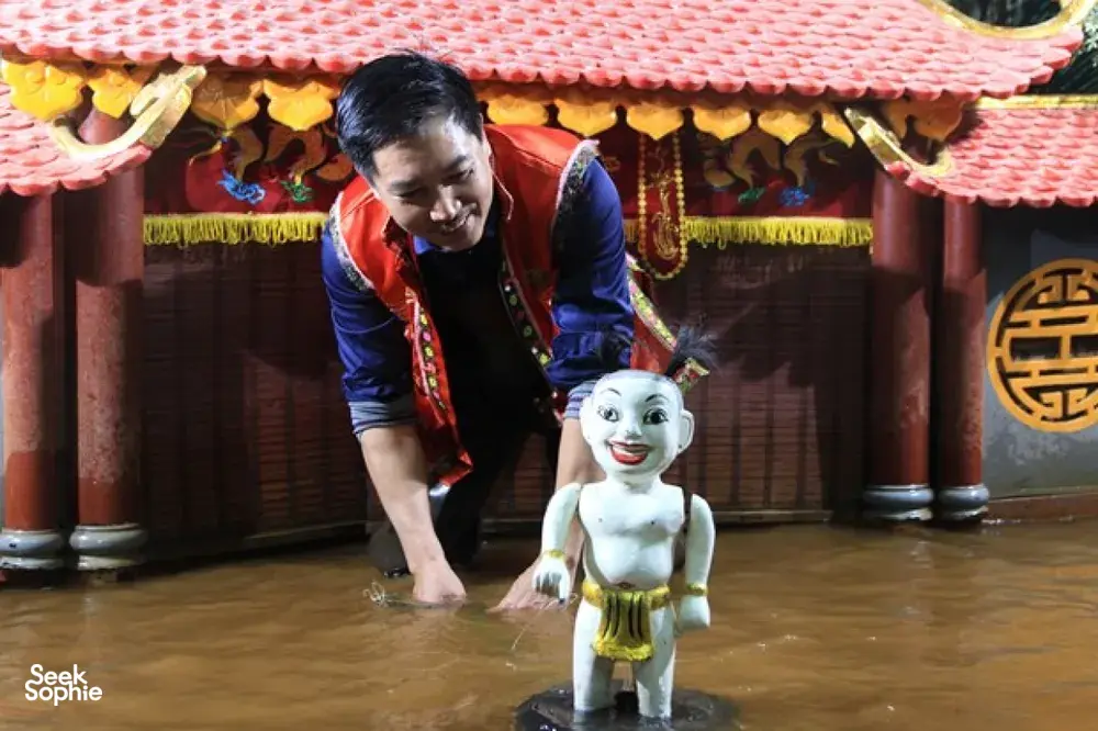一只迷你的白色水上木偶立在舞台地面上,操偶师正把它摆放在河内装饰精美的剧场里
