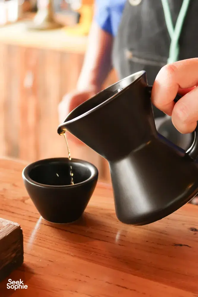 Hand-pouring dark coffee from a metal jug into a small cup during a specialty Yunnan coffee tasting in Lijiang