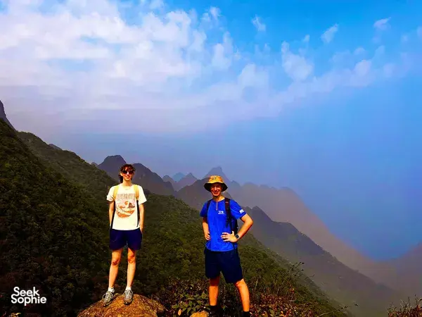 Two hikers pose on a ridge with layered mountain silhouettes behind them on Mt Fansipan in Sapa during a 1-day eco trek