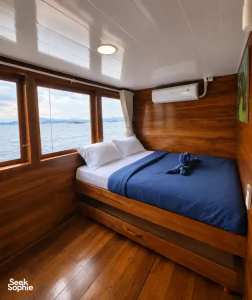 Wooden phinisi cabin with a double bed and sea view windows on a shared Komodo cruise, sailing from Labuan Bajo