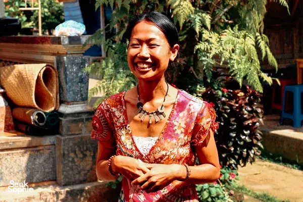 Smiling woman standing in a lush outdoor garden in Indonesia, wearing a red patterned dress and a beaded necklace in warm light