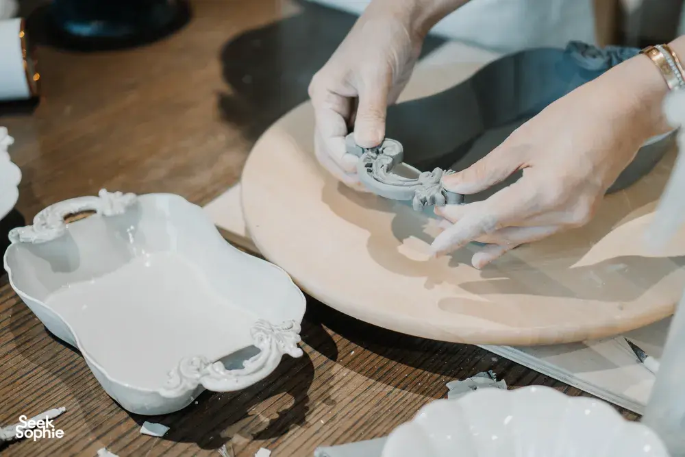 Ina Atelier potter’s hands fitting embossed ceramic pieces onto a plate base on the workbench at pottery workshops in Singapore