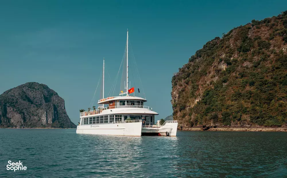 White sailing cruise yacht gliding on calm water with steep jungle-covered cliffs and a second vessel nearby, Halong Bay