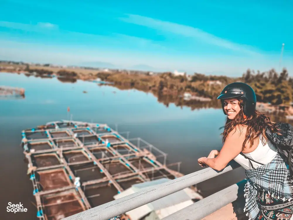 Helmed traveller smiling at sunrise on a riverside bridge in Hoi An, with long fishing boats moored in calm water