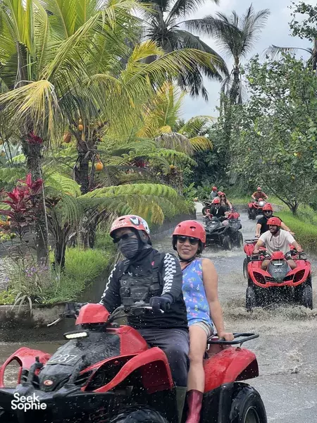 Riders in helmets on red ATV quad bikes on a Bali countryside track, surrounded by palm trees and greenery