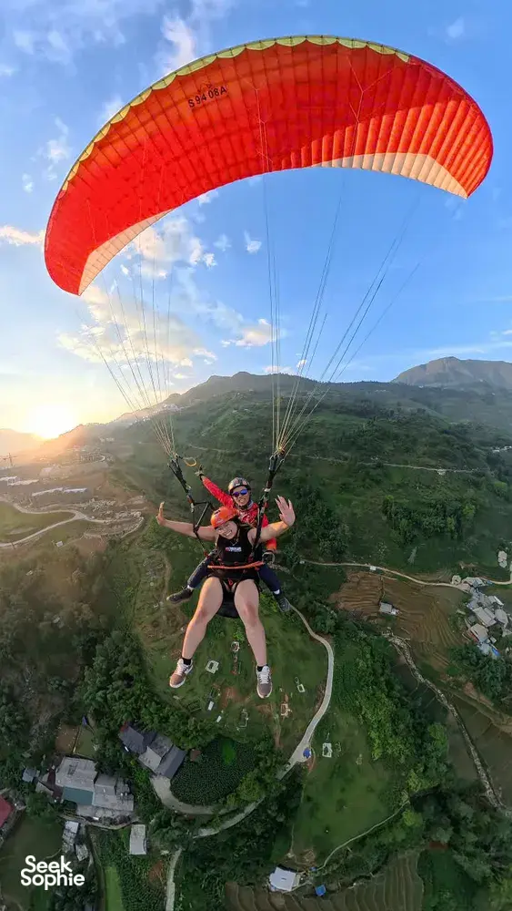 A paraglider duo flies beneath a bright red canopy over winding roads and villages during golden hour in Sapa