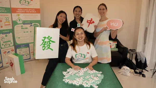 Team members pose behind a mahjong table in Singapore, holding character cards as the tiles are arranged for team building Singapore