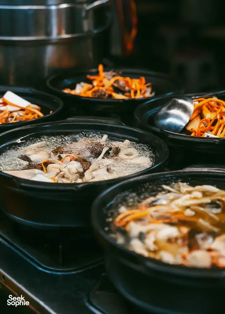 Rows of black soup pots at a Lijiang food stall with bubbling broth, fish pieces, and orange shredded vegetable topping