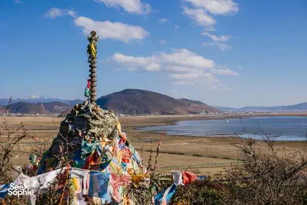 Colourful prayer-flag stone cairn overlooking a wide winter plain and lake in Shangri-La, with low mountains under bright sky